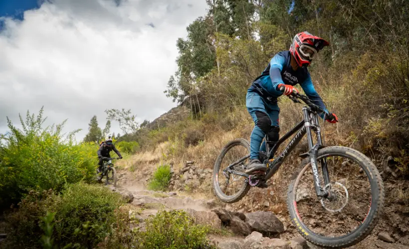 Mountain biker approaches a rocky feature on a downhill line in Lamay, Sacred Valley, Cusco, Peru, surrounded by dry shrubs.