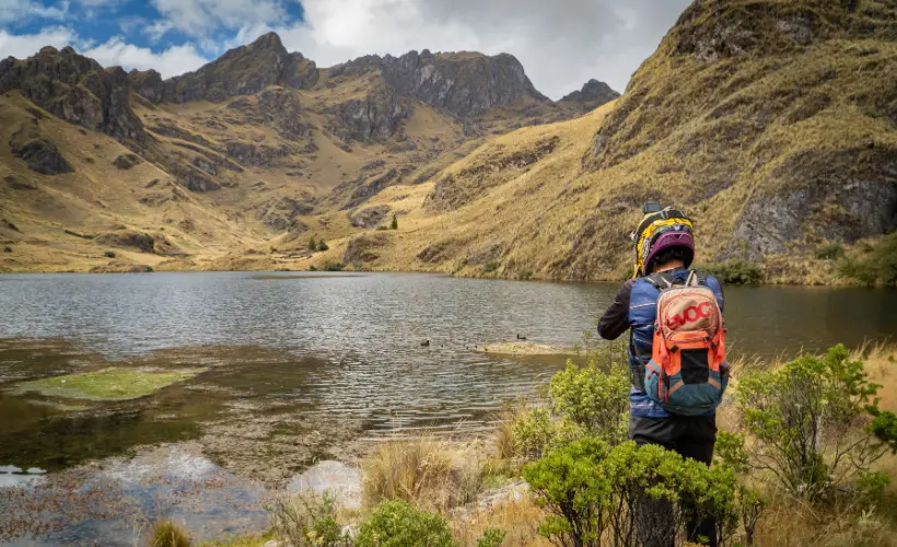 Ciclista con casco y mochila observa una laguna altoandina cerca de Lamay, Cusco, Perú, rodeada de lomas doradas y montañas.
