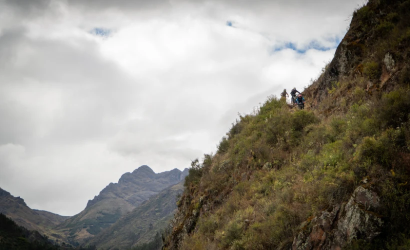 Cyclists traverse a cliffside path near Lamay in the Sacred Valley, Cusco, Peru, with rugged peaks, dry grass, and overcast skies.