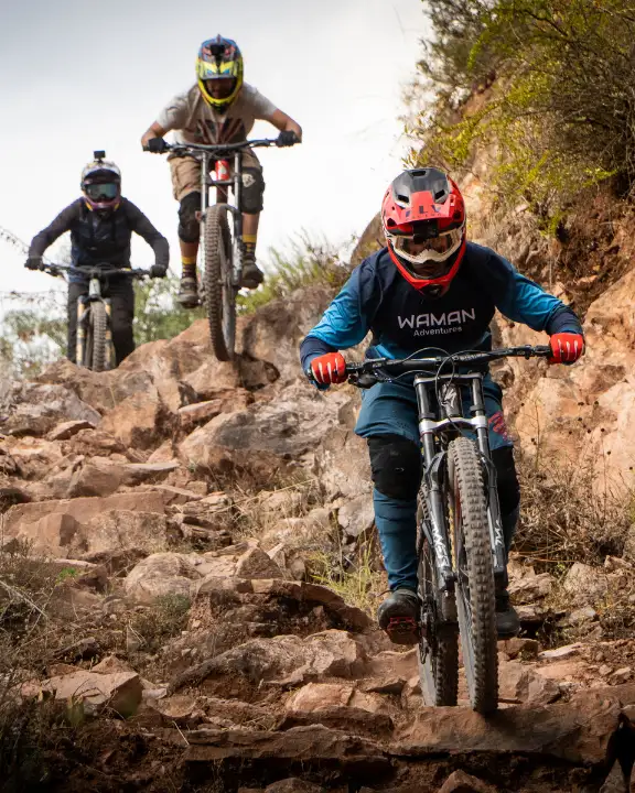 Tres ciclistas de montaña eligen líneas en un rock garden empinado en Lamay, Valle Sagrado, Cusco, Perú, levantando polvo y piedras.