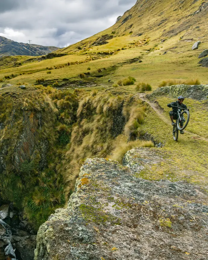 Un rider de downhill avanza por un sendero de alta montaña, rodeado de pastizales andinos y un profundo barranco natural.