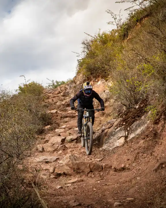 Rider de downhill bajando una canaleta con rocas sueltas en Lamay, Valle Sagrado cerca de Cusco, Perú, con casco integral, polvo y piedras.