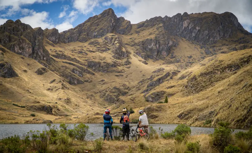 Grupo de ciclistas descansando junto a una laguna de altura en Lamay, Valle Sagrado, Cusco, Perú, con bicicletas, agua y picos montañosos.