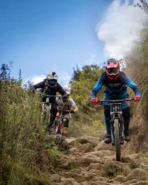 Riders de MTB pasando un rock garden técnico en Lamay, Valle Sagrado, Cusco, Perú, con cascos, piedras sueltas, ichu seco y montañas.