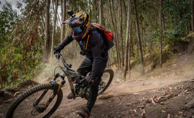 Mountain biker descending a loose trail through trees in Lamay, Sacred Valley, Cusco, Peru, wearing helmet, with dusty path and forest shade.