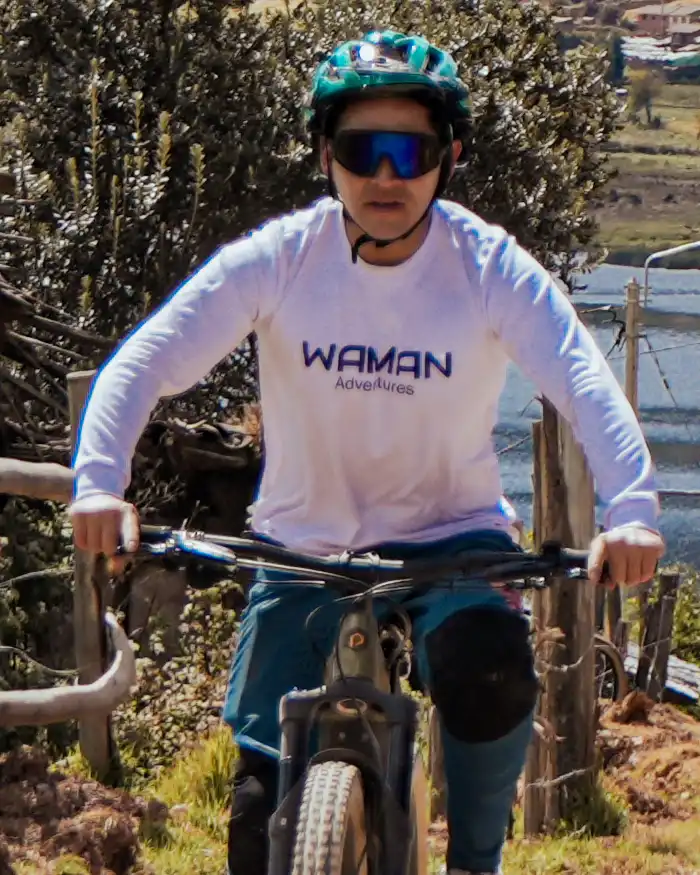 Mountain biker rides a community route in Cusco, Peru, with a calm lagoon behind and rugged Andean mountains along the trail.