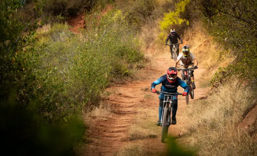 Rider train descending a tight singletrack ribbon in Lamay, Sacred Valley near Cusco, Peru, surrounded by shrubs, dust, and narrow trail edges.
