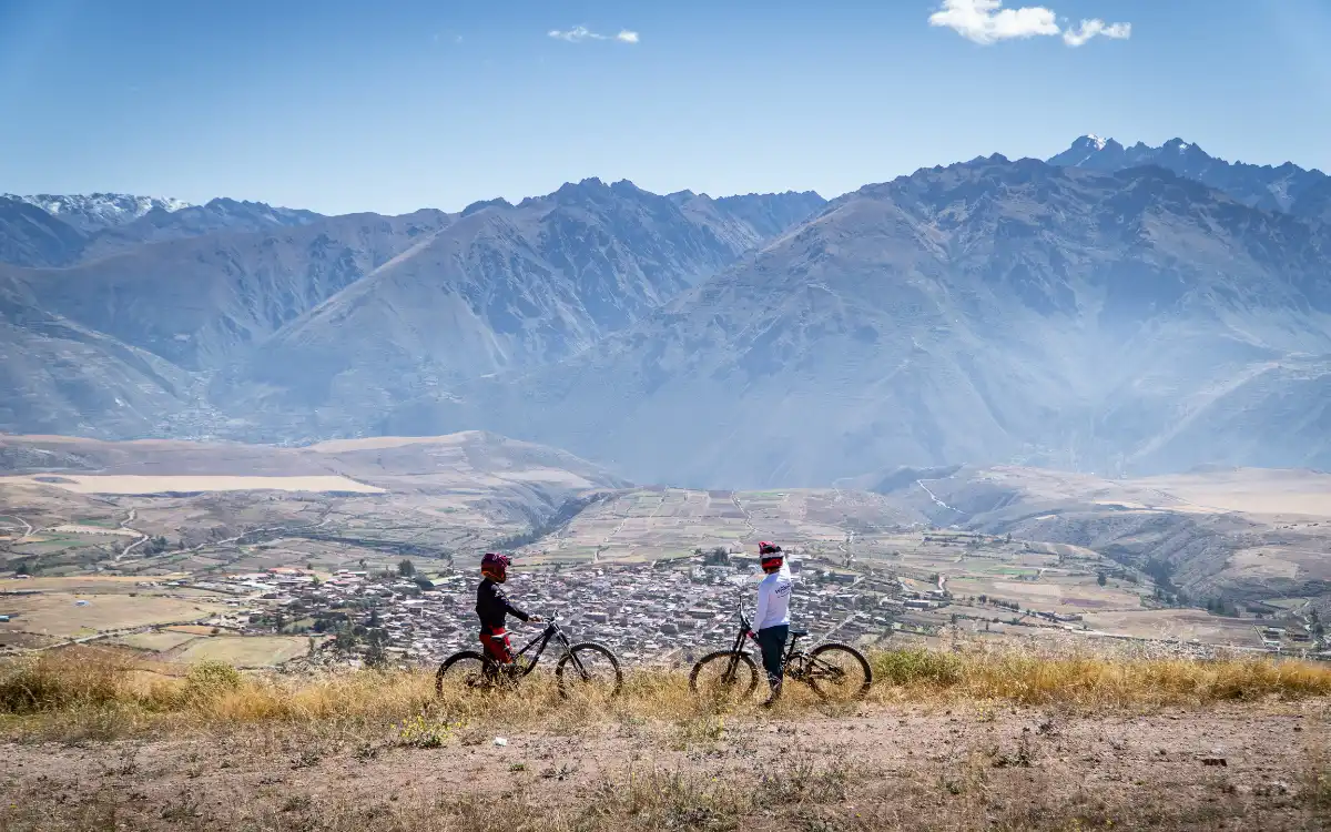 Two mountain bikers with bikes looking over the Maras valley panorama in the Sacred Valley, on the Epic MTB Maras Trail, with distant peaks and clear sky.