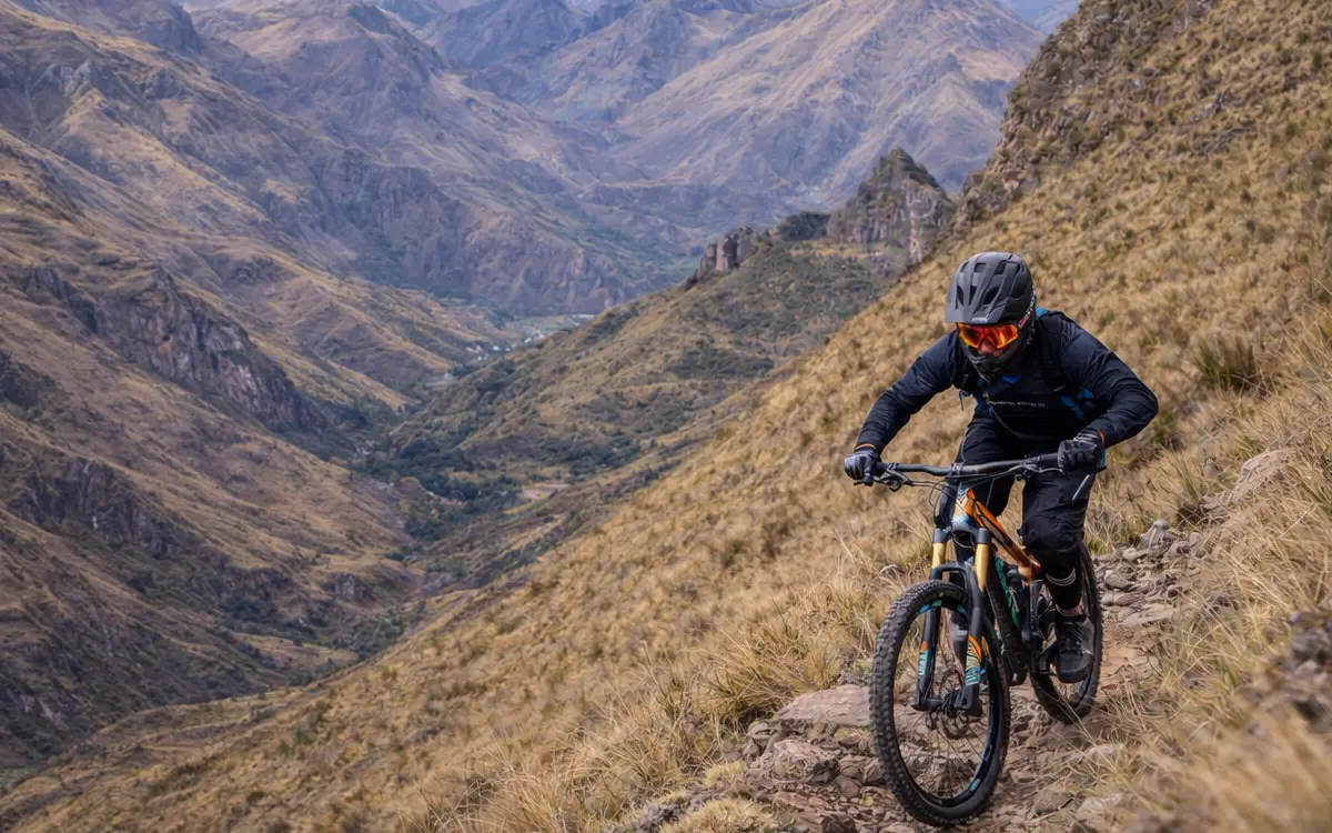 Mountain biker riding a narrow dirt singletrack above a deep valley, with wide Andean mountains and dry grass slopes.