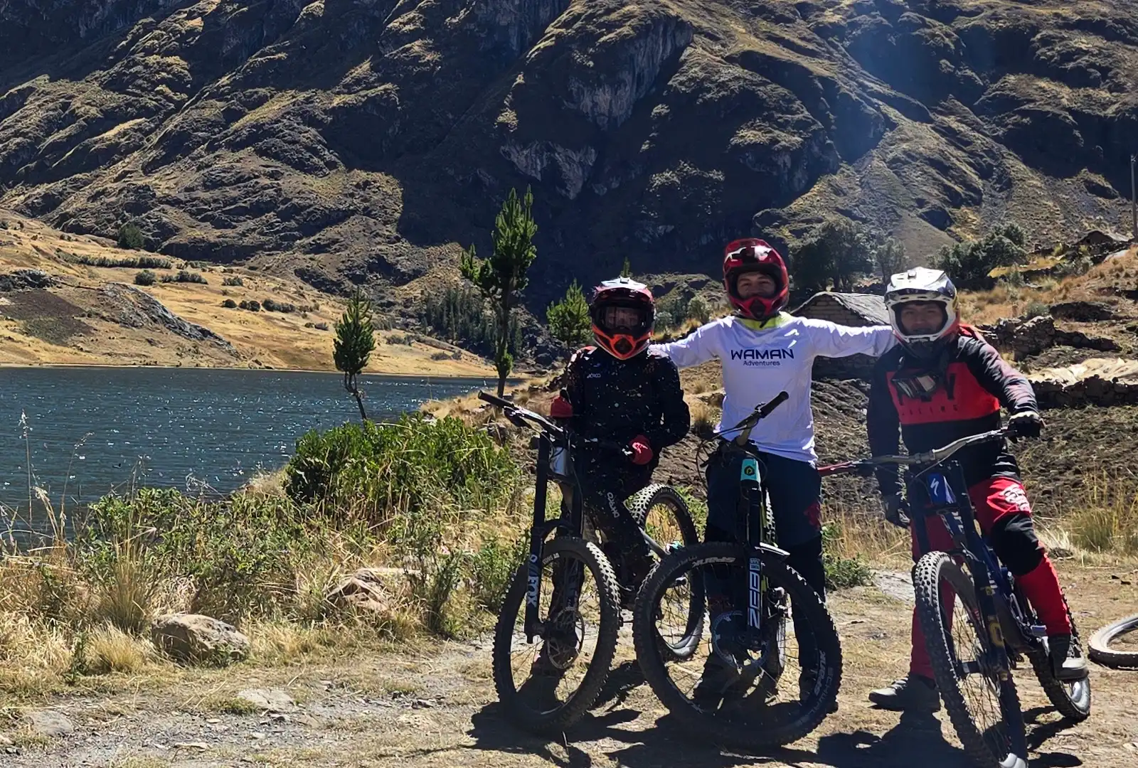 Tres ciclistas de montaña posan junto a una laguna andina, en una pausa del recorrido. Downhill vs Enduro vs E-Bike en Cusco