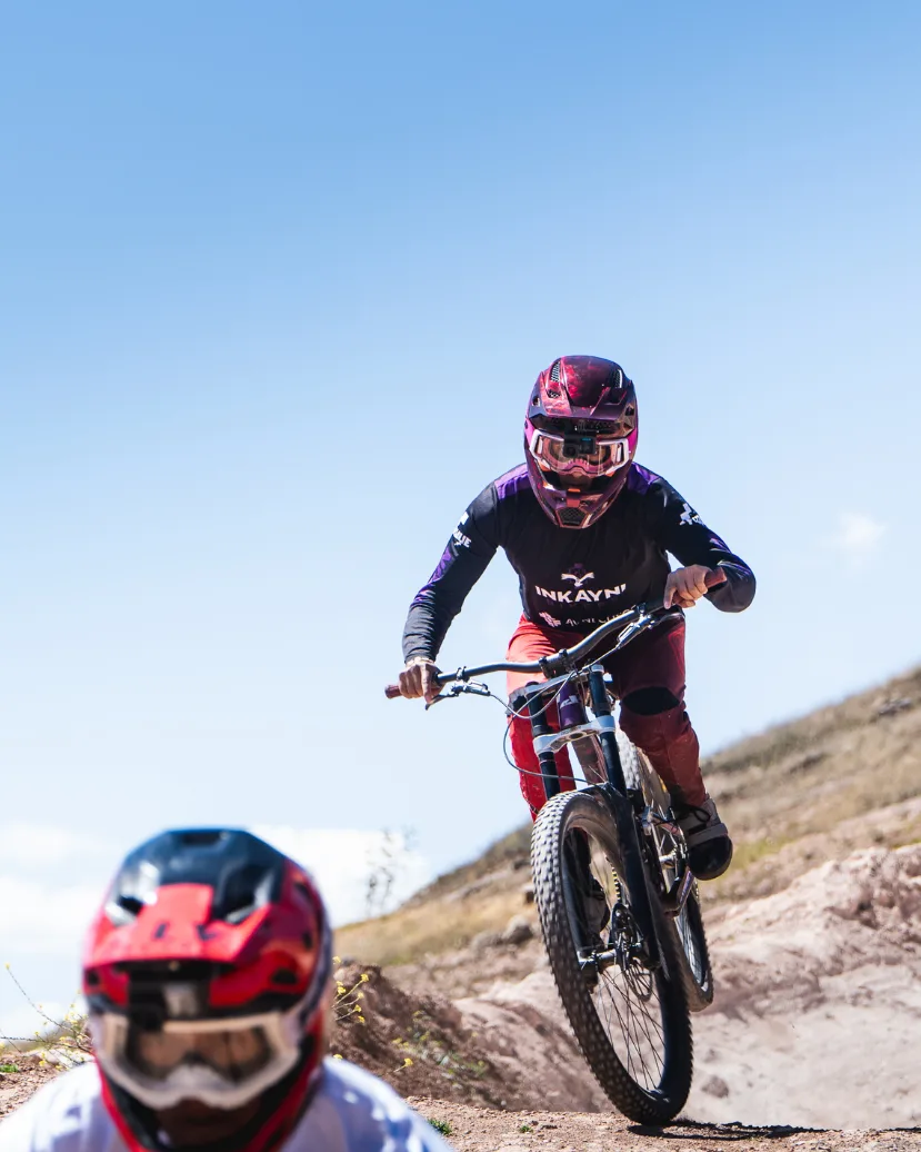 Two downhill mountain bikers dropping into a dusty Maras trail in the Sacred Valley near Cusco, Peru, wearing full face helmets and pads.