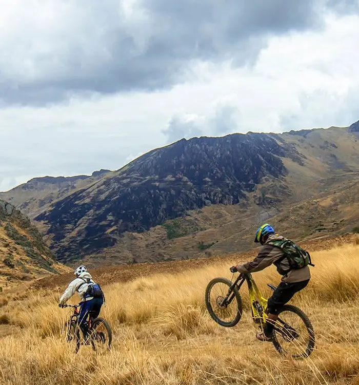 Dos ciclistas de montaña avanzan por pastizales abiertos de los Andes, rodeados de colinas y montañas.