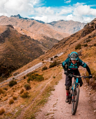 Fotografía de ciclista de montaña descendiendo por camino de tierra en Valle Sagrado de los Incas, vistiendo casco turquesa y mochila, sobre bicicleta turquesa, con montañas áridas marrones del altiplano cusqueño al fondo bajo cielo azul con nubes blancas