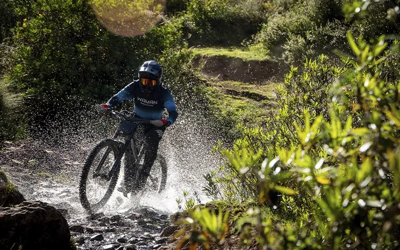 A cyclist practicing mountain biking in cusco crosses a stream, splashing water on a trail surrounded by lush vegetation