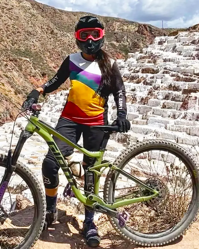 An enduro rider poses with her mountain bike in front of the Maras salt ponds under a deep blue sky.