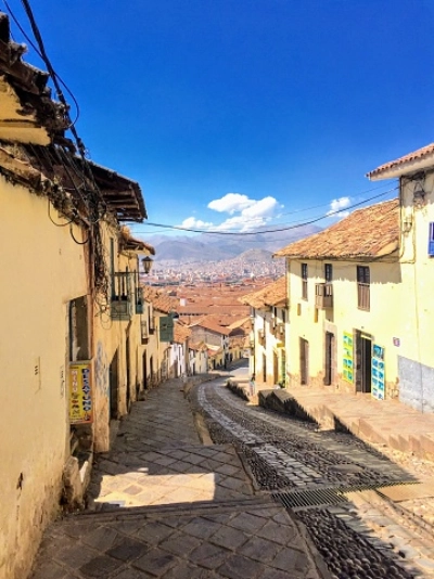 Steep downhill street in the Santa Ana neighborhood of Cusco with stone paving and a wide view of the city rooftops under a bright blue sky.
