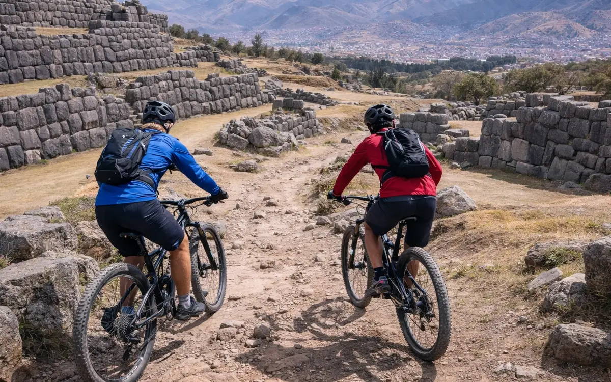 Two mountain bikers riding dirt and rocky trails near Sacsayhuaman in Cusco, Peru, with Inca stone walls and wide valley views around.