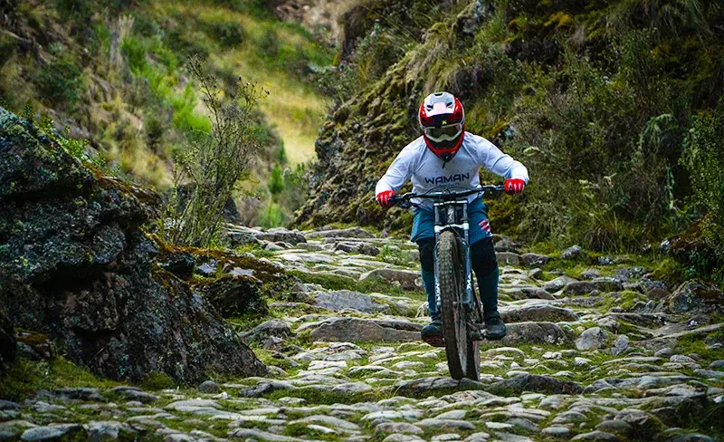 Mountain biker wearing a red helmet and Waman Adventures shirt descends a rugged stone path riding mountain bikes on Inca stairs in Cusco.