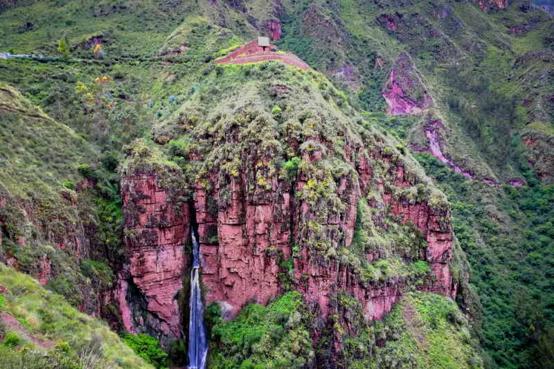 Panoramic view of the catarata de Perolniyoc flowing down reddish cliffs below the Raqaypata ruins on the Perolniyoc trail route in Cusco.