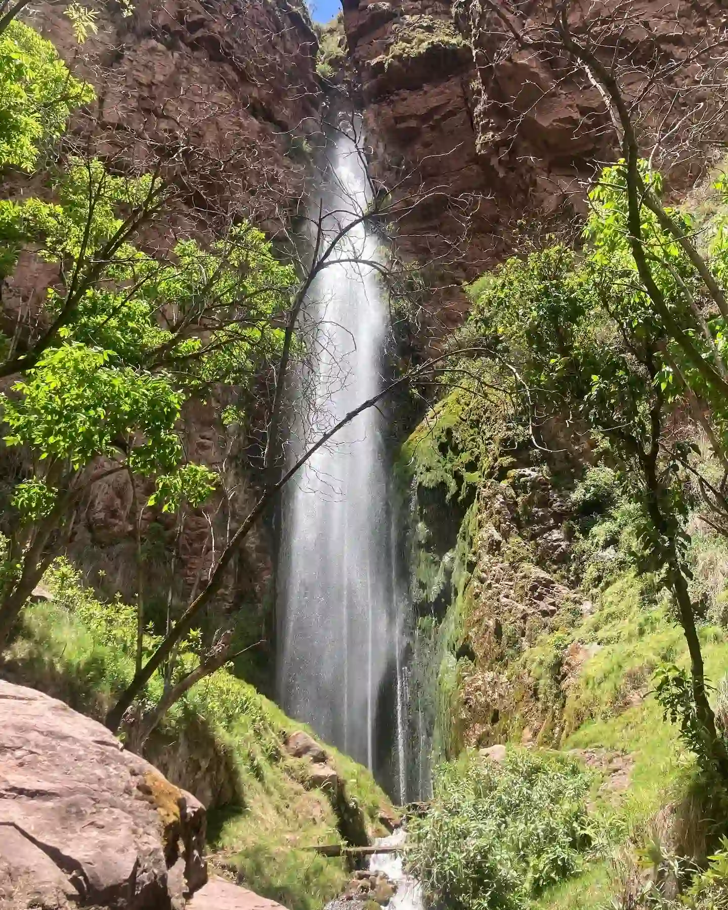 Tall waterfall flowing between high reddish rock walls and lush green vegetation illuminated by the sun at the Perolniyoc waterfall in Perolniyoc trail in Cusco