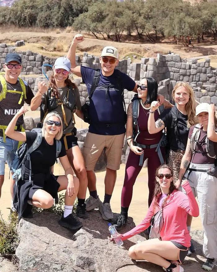 Group of visitors at Sacsayhuaman in Cusco, Peru, with massive Inca stone walls behind them, wearing backpacks and casual travel clothing.