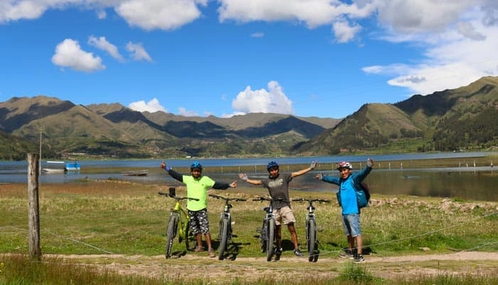 Three cyclists pose next to their bikes in front of a lake and mountains under a blue sky, along the Piuray Chinchero & Huaypo route during a E-bike in Cusco experience