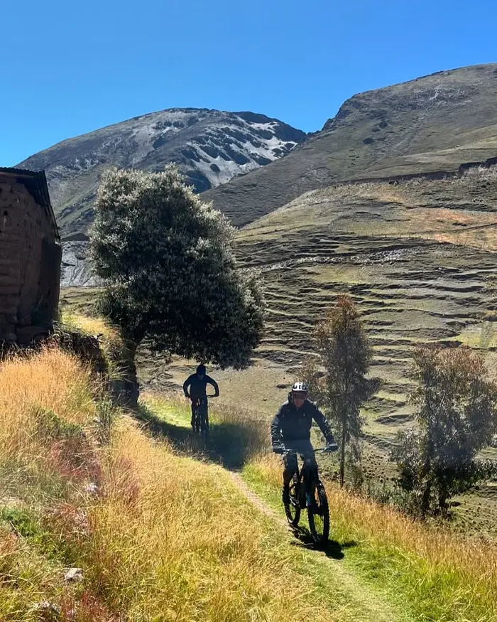 Dos ciclistas avanzan por un sendero angosto cubierto de pasto verde que corta la ladera dorada de la montaña.