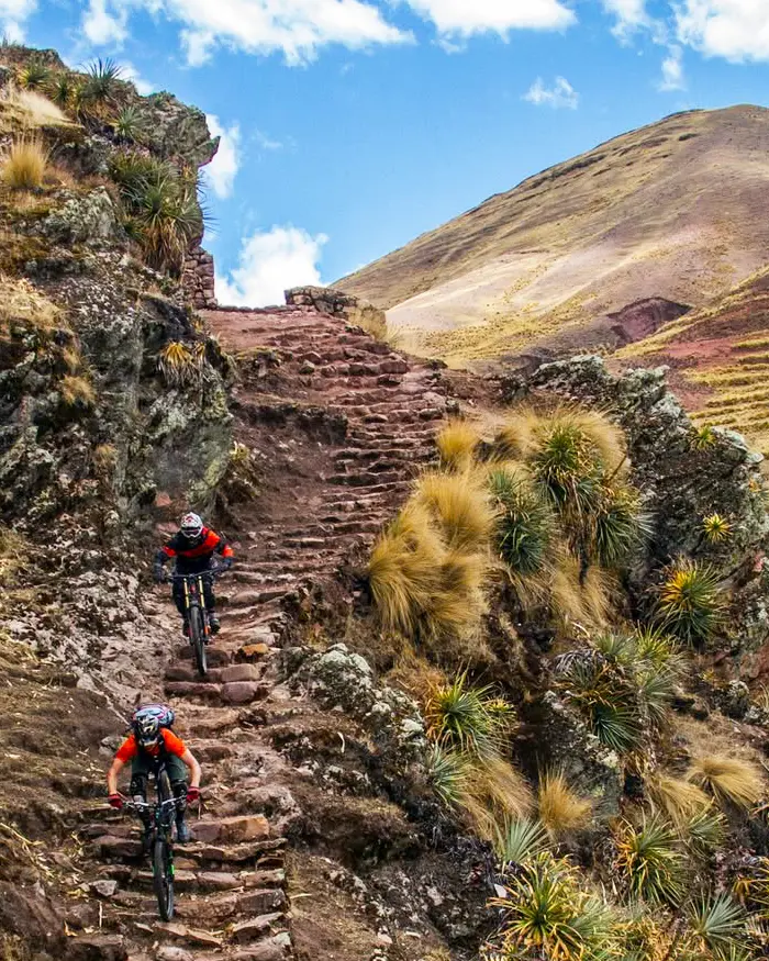 Two mountain bikers descending steep stone steps in Cusco, Peru, with rock walls, dry grass, and a narrow technical line.