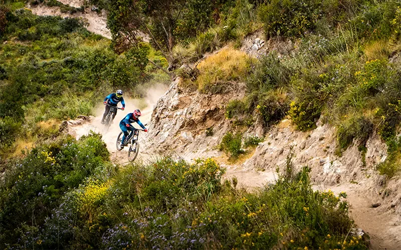 Two mountain bikers descend rapidly down a narrow, dusty dirt trail, kicking up significant dust while practicing Enduro en Cusco on the Perolniyoc trail.