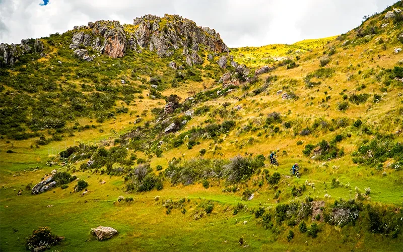 Two mountain bikers riding down a dirt path among green, rocky hills under clouds while doing enduro in Cusco on the Perolniyoc Trail.