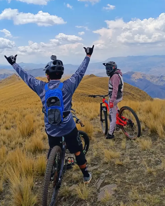 Two mountain bikers resting on a high Andean summit near Cusco, Peru, with golden ichu grass, open sky, and mountain views.