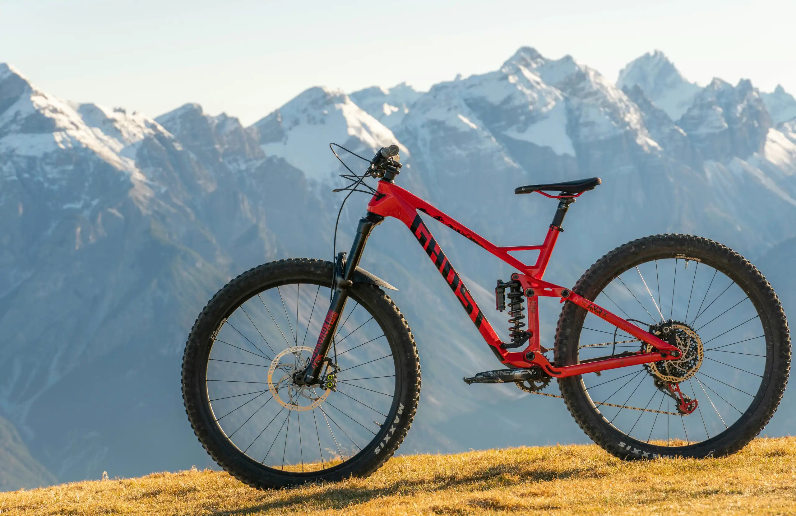 Red full-suspension enduro mountain bike parked on grass with dramatic snowcapped mountains in the background.