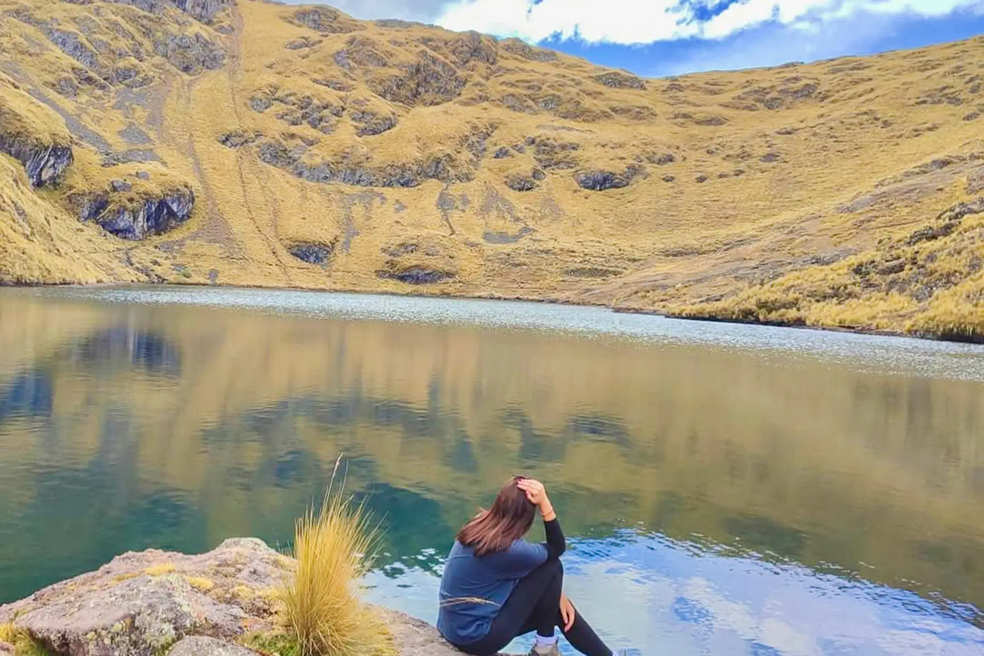 Viajera sentada sobre una roca junto a la laguna Azulcocha en las alturas de Pisac, con el agua en calma reflejando laderas doradas andinas.