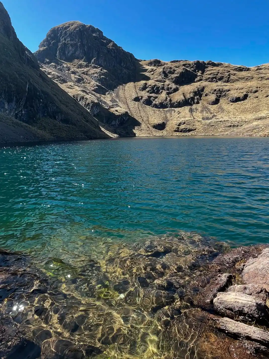 Laguna altoandina de agua azul y transparente con orilla rocosa y montañas andinas bajo un cielo despejado en las alturas de Pisac.