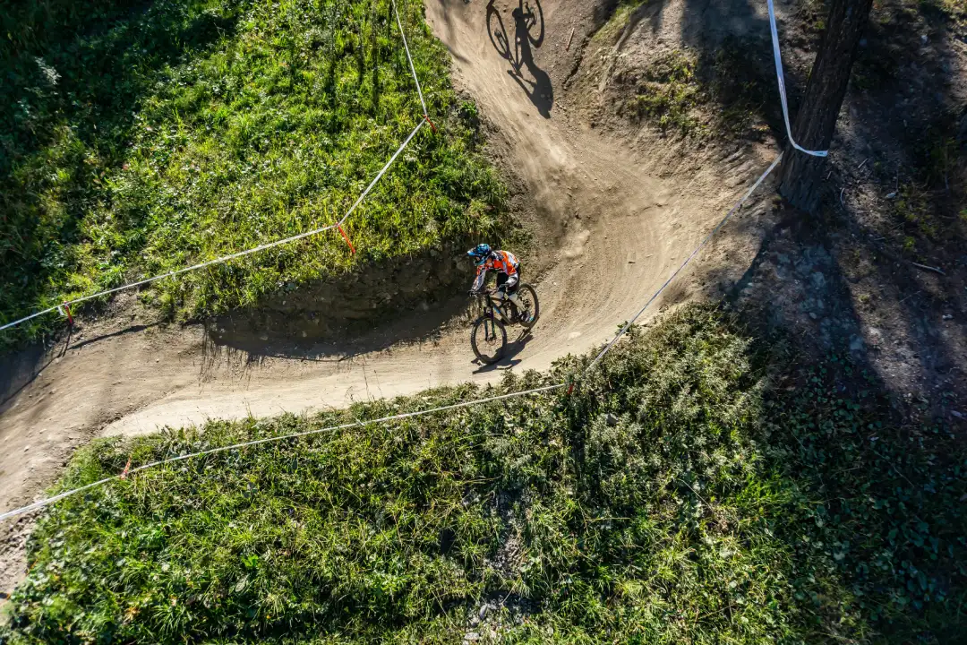 Overhead view of a mountain biker leaning through a tight switchback berm on a dirt trail, surrounded by green grass and trees.