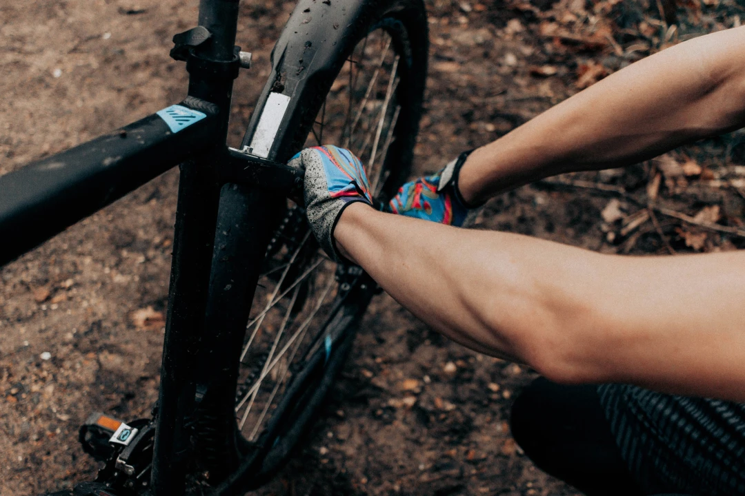 Close-up of a rider’s hands adjusting a mountain bike wheel on a dirt trail, showing gloves, frame, and tire details.