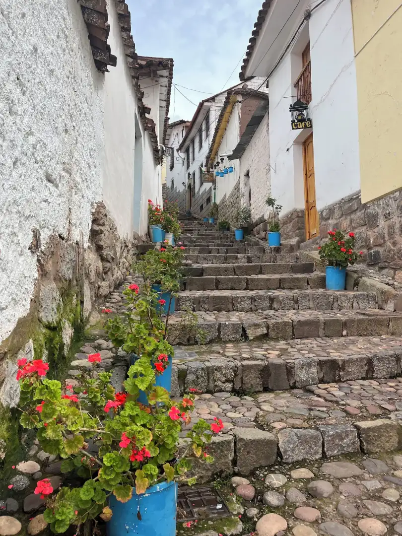 Narrow Cusco street with uneven stone steps and cobblestones, lined with blue flower pots, which can be slippery when wet.