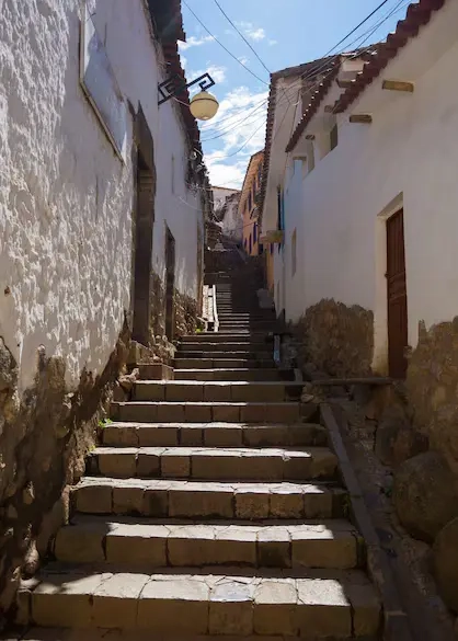 Steep stone stairway in a narrow alley in San Blas, Cusco, with white colonial walls and traditional rooftops under daylight. Cusco Urban Downhill