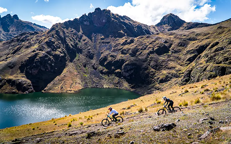Dos ciclistas de montaña rodando por un sendero altoandino sobre una laguna azul, con montañas escarpadas al fondo en las alturas de Pisac. las 3 Lagunas de Pisac en E-Bike