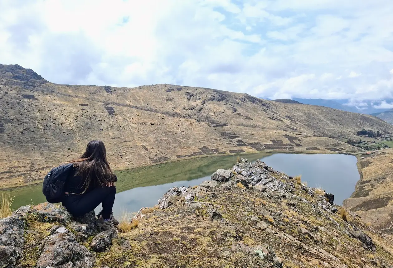 Viajera sentada sobre rocas con vista a la laguna Kinsacocha en las alturas de Pisac, con laderas amplias andinas y cielo nublado.