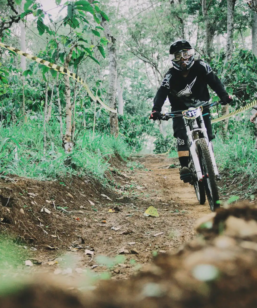 Downhill mountain biker wearing a full-face helmet riding on a loamy dirt trail through a green forest, with course tape along the track.