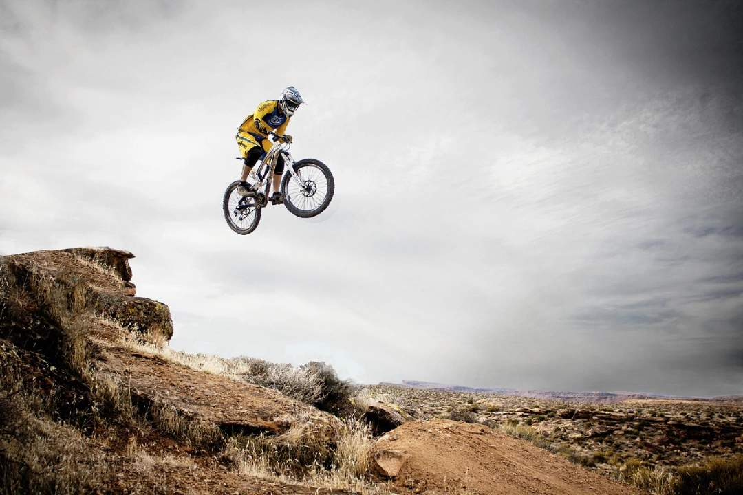 Spectacular photograph of freeride cyclist airborne executing jump from rock formation, wearing yellow jersey and full-face helmet, dual-suspension bike with wheels suspended in air, arid desert landscape in background, dramatic cloudy sky, advanced drop-off technique