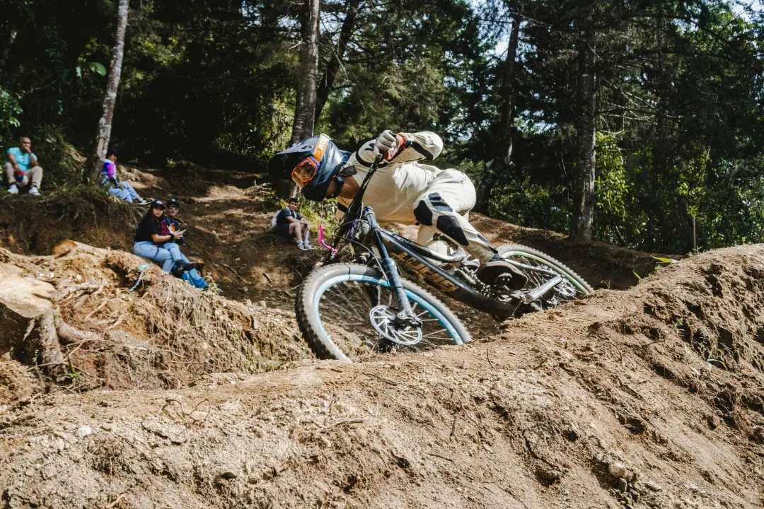 Fotografía de acción de ciclista profesional de downhill navegando curva técnica en terreno lodoso empinado, vistiendo equipo completo (casco integral con gafas naranja, traje beige de protección, guantes), bicicleta de doble suspensión azul y blanca, espectadores sentados observando en ladera de bosque al fondo