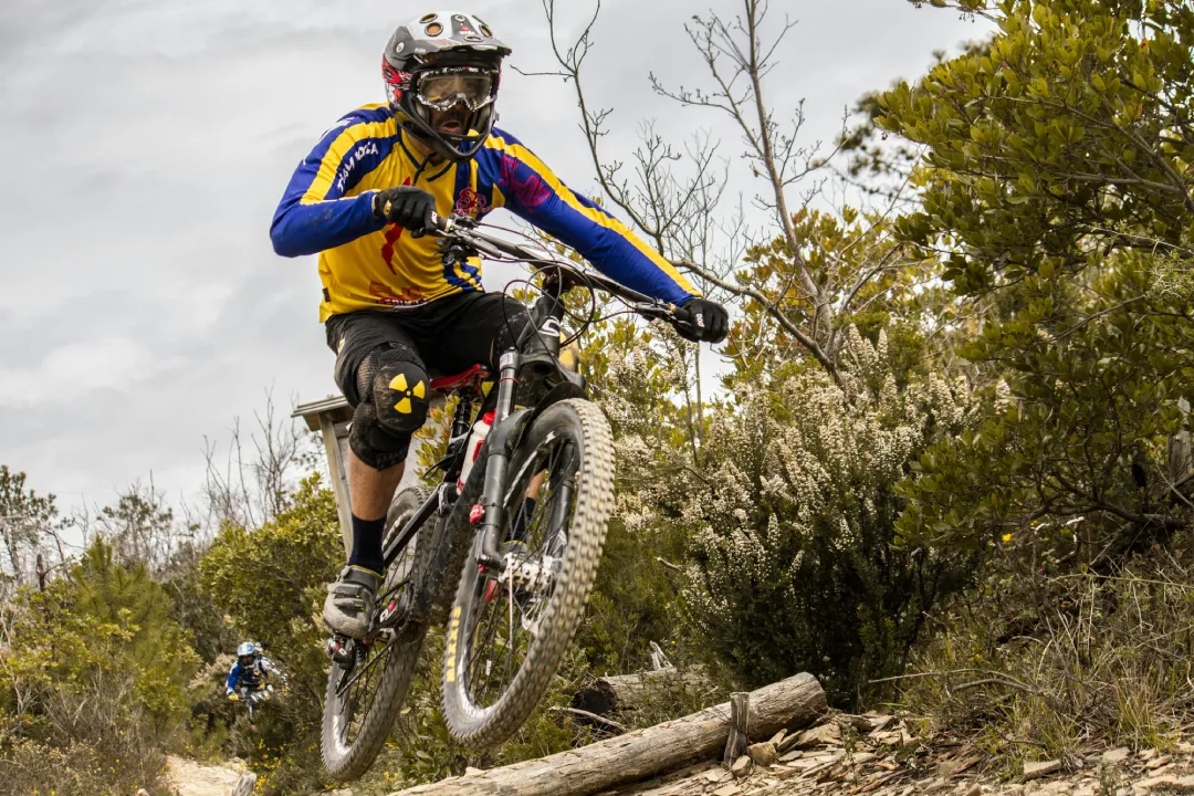 Downhill mountain biker wearing a full face helmet jumping over rocks on a rugged trail, with another rider in the background