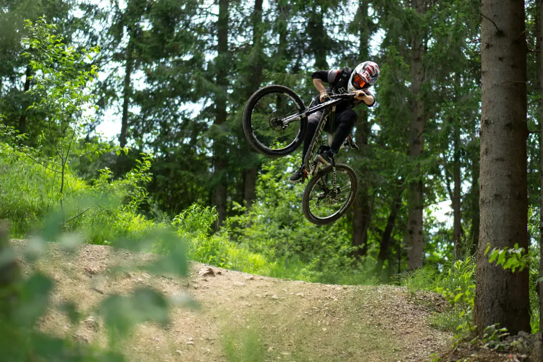 Mountain biker wearing a full-face helmet performing a high jump on a dirt trail in a green forest.