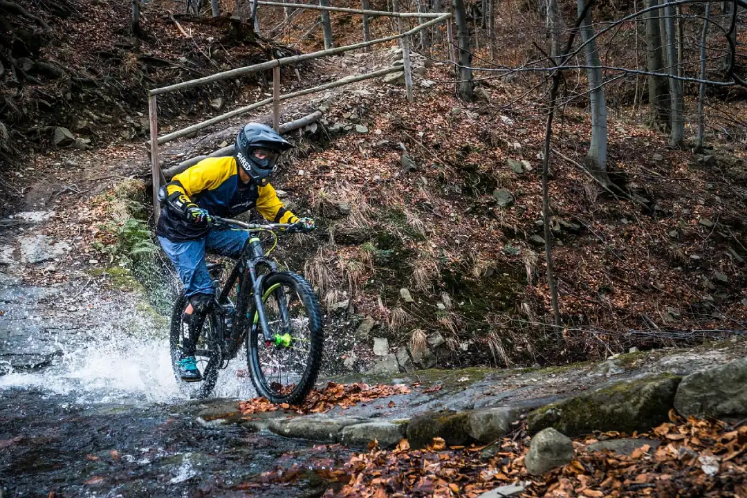 Action photograph of enduro cyclist crossing rocky stream with water splashes in autumn forest, wearing full gear (full-face helmet, yellow and blue jersey, neon gloves, blue pants), enduro bike with mud tires, brown dry leaves on ground, metal trail protection barriers in background, leafless trees