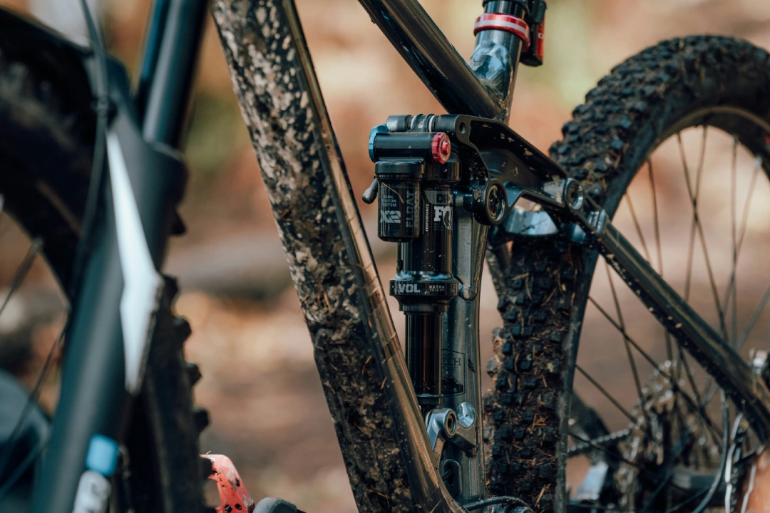 Close-up of a mountain bike rear suspension shock and linkage with muddy frame and tire, showing suspension components in detail.