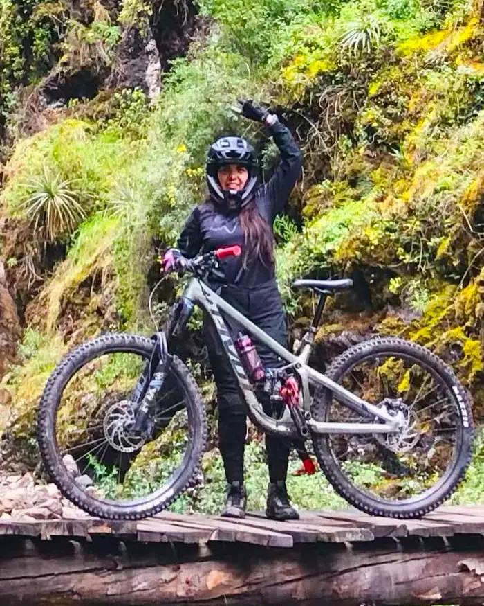A cyclist stops on a wooden bridge in the middle of a mountainous environment full of vegetation.