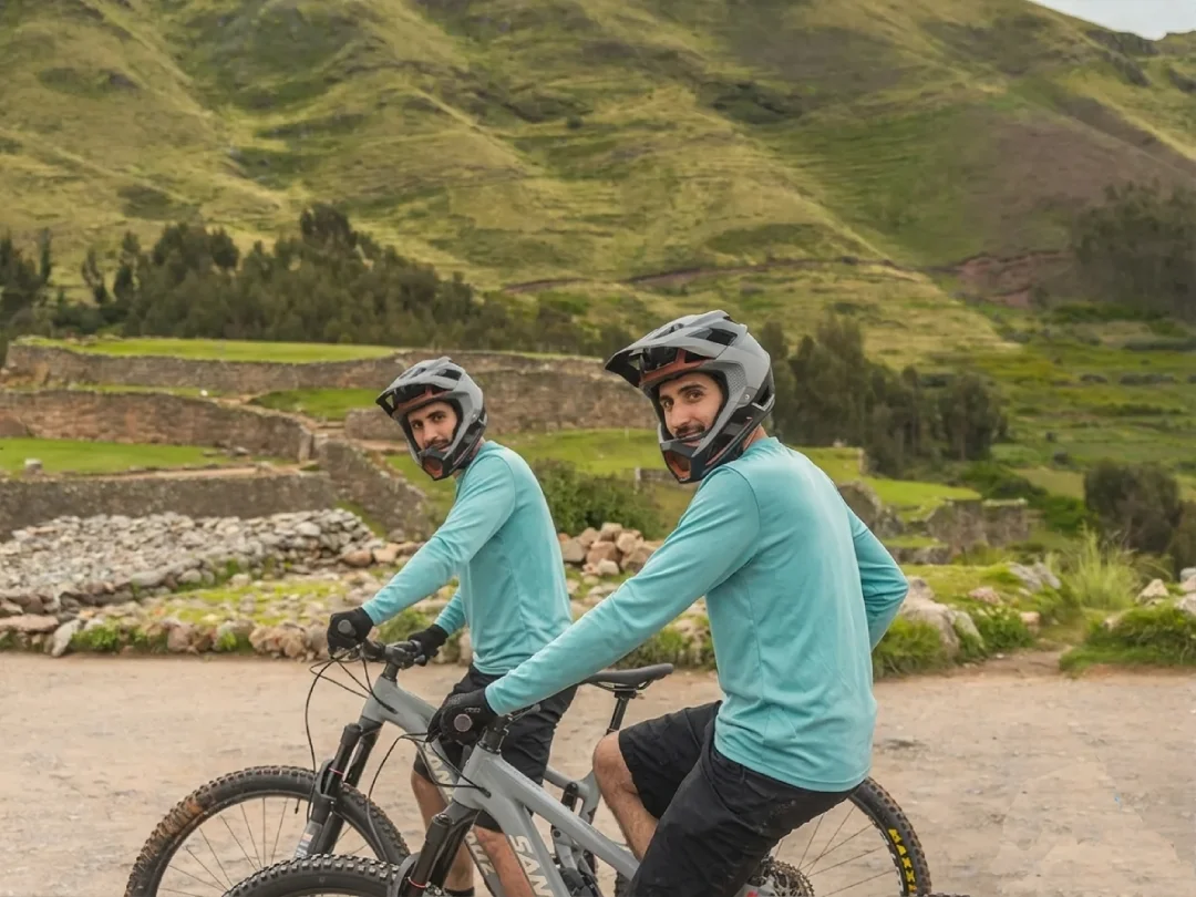 Two mountain bikers wearing helmets and matching blue jerseys pause on a dirt road with green Andean hills and stone terraces in the background.