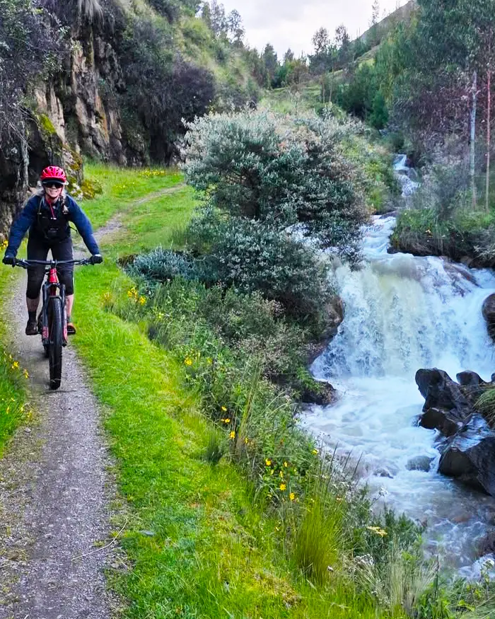 La ruta serpentea junto a una pequeña cascada, creando una escena fresca y llena de vida en el entorno andino.